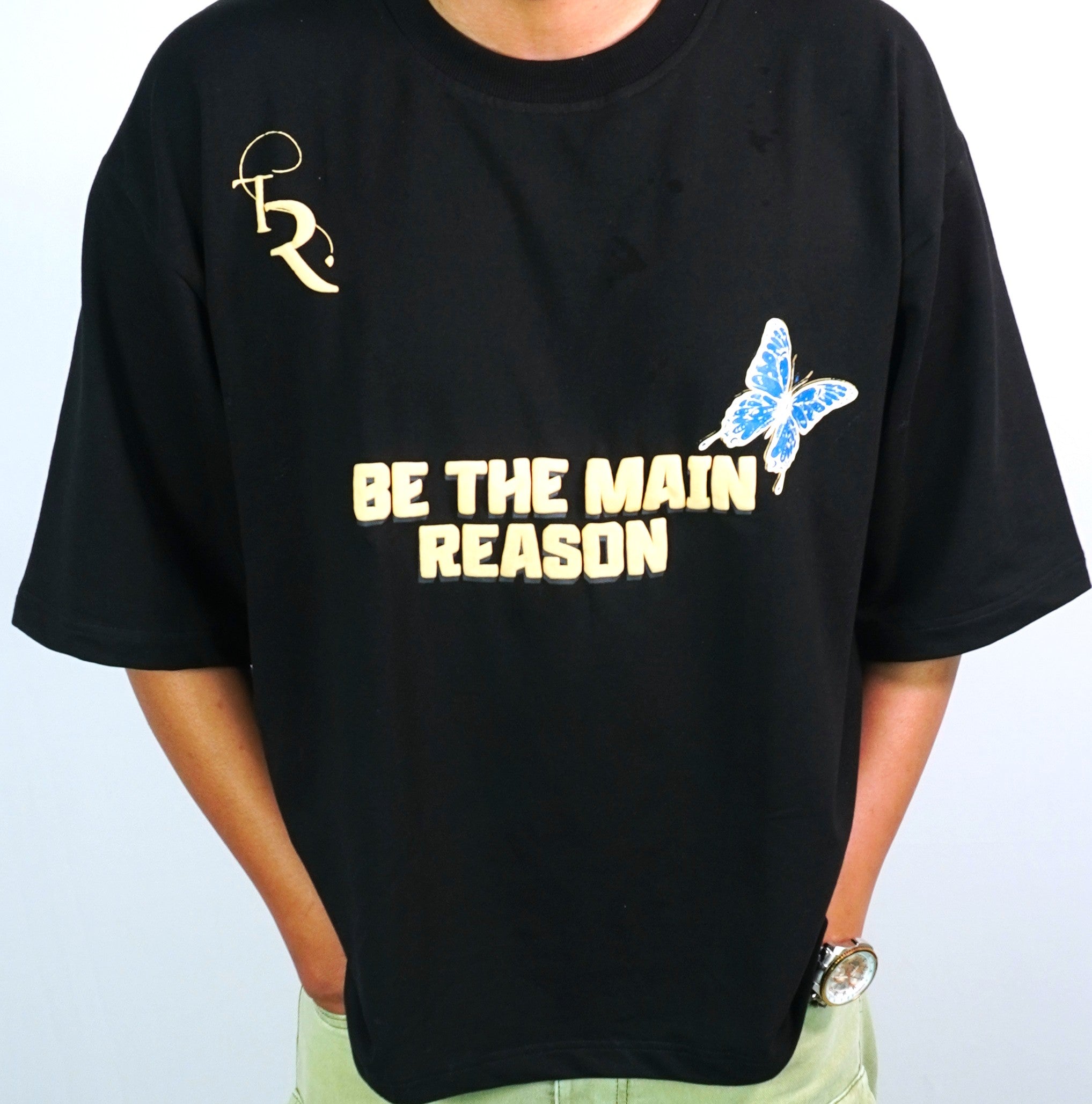 Close-up of TAMRAT black boxy fit t-shirt showing 'BE THE MAIN REASON' typography and blue butterfly chest print