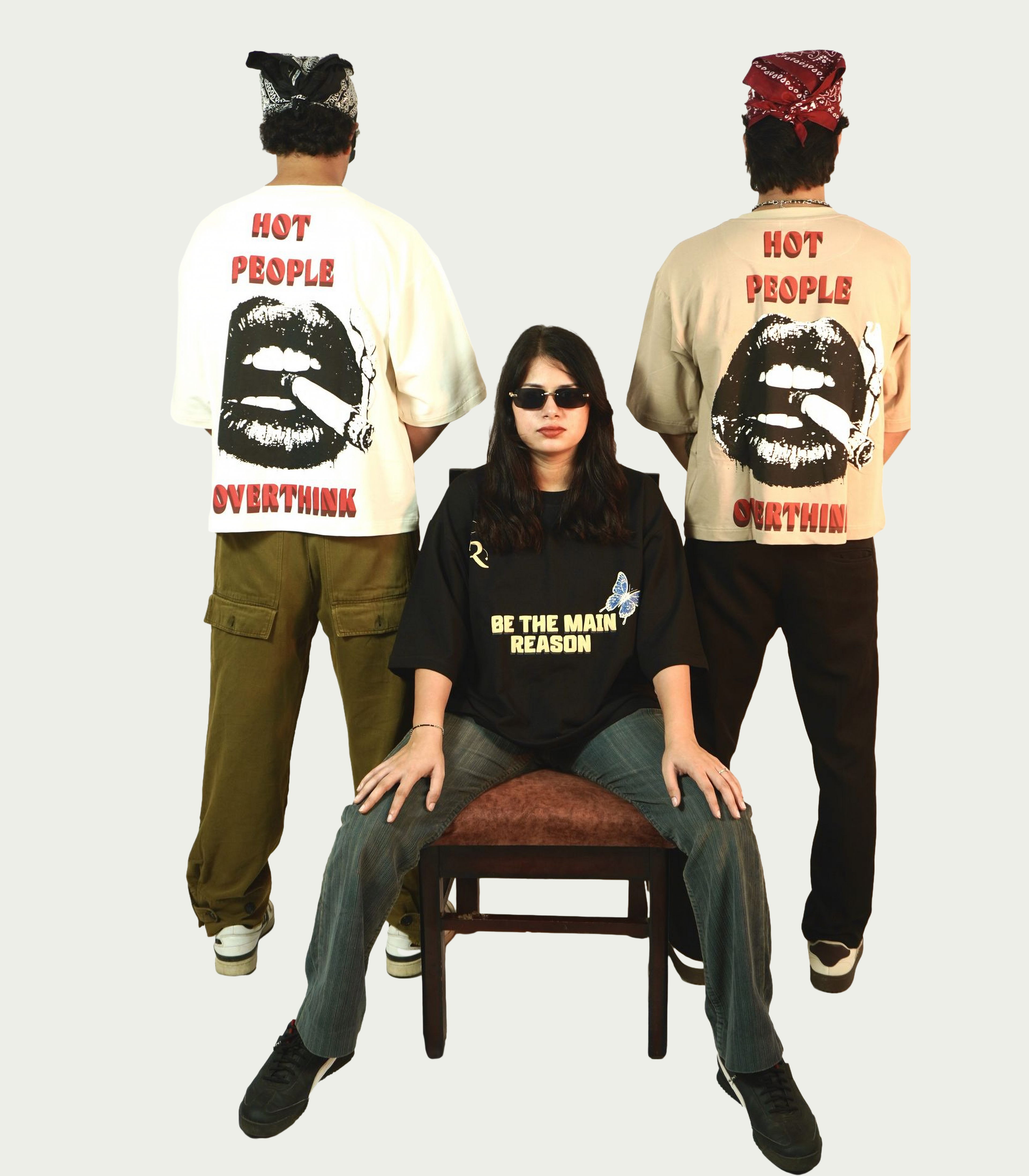 Three models shown from the back wearing graphic t-shirts with a large lip print and the text 'HOT PEOPLE OVERTHINK' on a light gray background. The model in the center sits on a small stool wearing a black t-shirt that reads 'BE THE MAIN REASON'.